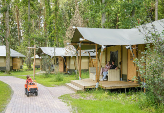 Safari tents in a wooded area, child driving a toy car, and couple relaxing on the porch outside.