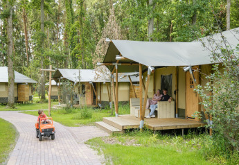 Tiendas de safari en el bosque con un niño conduciendo un coche de juguete y pareja en la terraza.