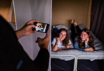 Twee meisjes liggen lachend op een bed in een safaritent terwijl iemand een foto maakt met een telefoon.