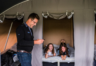 Un homme sourit debout près de deux filles riant, allongées sur un lit dans une tente safari.
