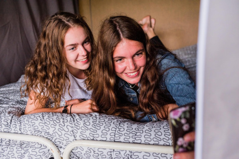 Deux jeunes femmes sur un lit dans une tente safari prennent un selfie en souriant au Resort Echternacherbrück en Allemagne.