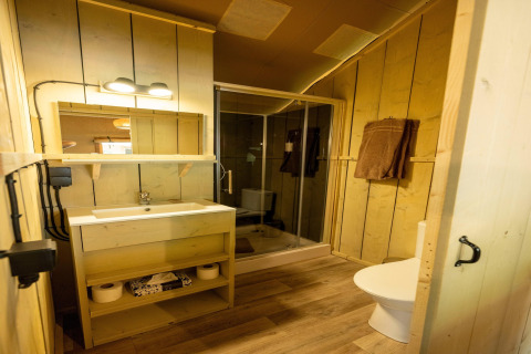 Cozy safari tent bathroom with wooden walls, a shower enclosure, modern sink, towels, and a toilet.