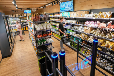 Shopping area in a safari tent with people, shelves of snacks, plush toys, and security monitors.