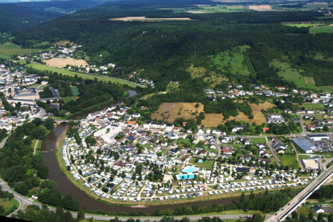 Luchtbeeld van een camping aan een rivier vlak bij een dorp, omlijst door groene heuvels en velden.