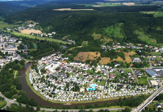 Vue aérienne d’un camping en bord de rivière près d’une ville, entouré de forêts, collines et champs.