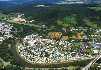 Luchtfoto van een camping aan een rivier bij een dorp, omringd door bossen, heuvels en akkers.