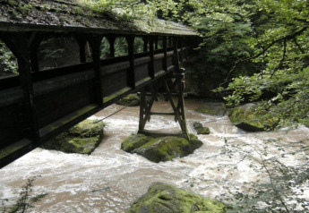 Overdekte houten brug over kolkende rivier, omringd door met mos begroeide rotsen en weelderig groen bos.