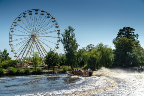 A large Ferris wheel and a group riding a speedboat on the river, surrounded by lush green trees.