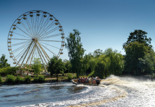 A large Ferris wheel and a group riding a speedboat on the river, surrounded by lush green trees.