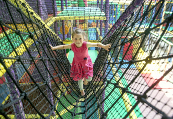 Een meisje in een roze jurk speelt op een touwbrug in een kleurrijke indoorspeeltuin met safari thema.