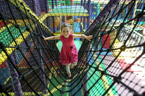 A child in a pink dress walks on a rope bridge inside a colorful indoor play area with a safari theme.