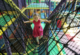 A child in a pink dress walks on a rope bridge inside a colorful indoor play area with a safari theme.