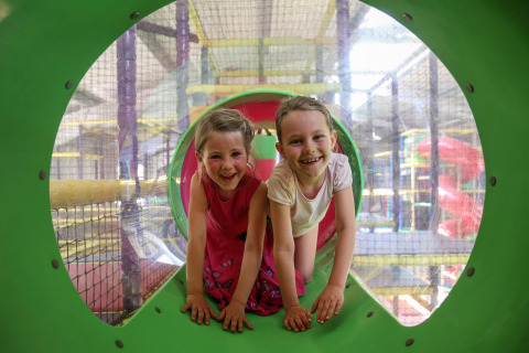 Two happy children playing inside an indoor play center, smiling while crawling in a green tunnel.
