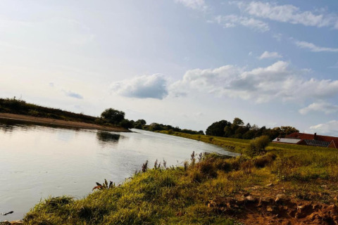 View from a safari tent area showing a river, grassy bank, trees, and a house under a cloudy blue sky.