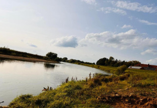 View from a safari tent area showing a river, grassy bank, trees, and a house under a cloudy blue sky.