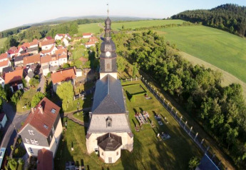 Luchtfoto van een Duits dorp met kerk, klokkentoren, groene velden en huizen met rode daken.