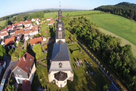Aerial view of a village in Germany with a church, clock tower, green fields, and red-roofed houses.