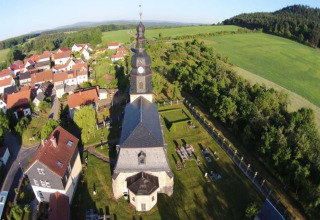 Aerial view of a village in Germany with a church, clock tower, green fields, and red-roofed houses.