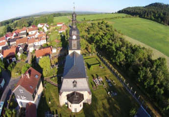 Aerial view of a village in Germany with a church, clock tower, green fields, and red-roofed houses.
