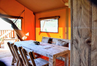 Interior of a safari tent with a rustic wooden table, chairs, orange canvas walls, and a window in daylight.