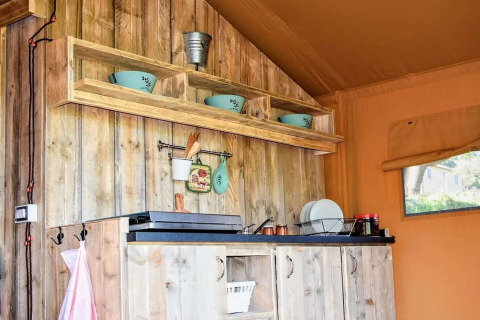 Rustic kitchen inside a safari tent with wooden cabinets, open shelving, blue bowls, and a side window.