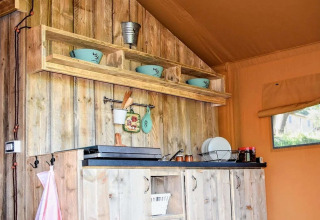 Rustic kitchen inside a safari tent with wooden cabinets, open shelving, blue bowls, and a side window.