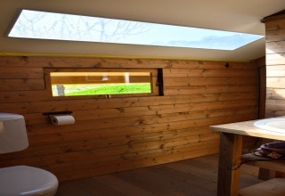 Safari tent bathroom with wooden walls, skylight, window, toilet, and sink, featuring natural light.