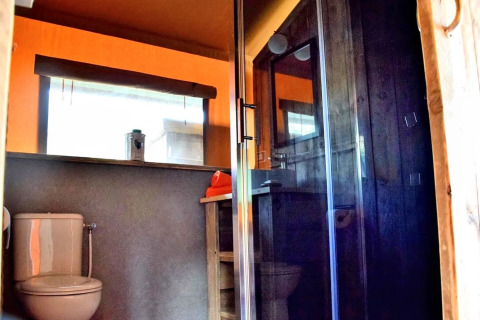 Bathroom inside a safari tent featuring a toilet, glass shower enclosure and a large window with sunlight.