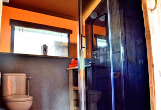 Baño dentro de una tienda safari con inodoro, ducha de vidrio, detalles en madera y ventana luminosa.