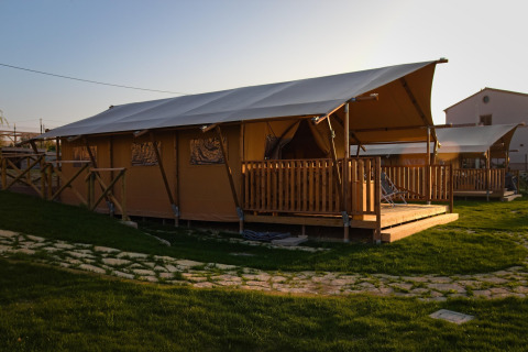Safari tent 'Tenda 1' in Italië bij zonsondergang, met houten terras en groene grasvelden eromheen.