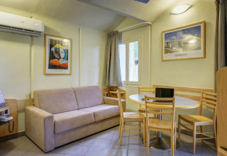 Mobile home living area with beige sofa, round dining table, wooden chairs, wall art, and air conditioning.
