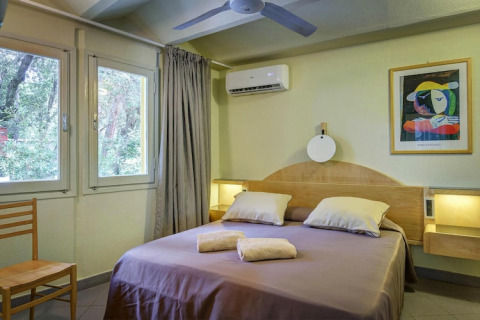 Schlafzimmer in einem Mobilheim mit Doppelbett, Fenstern, Klimaanlage, Stuhl und bunter Wandkunst.