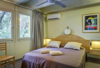 Schlafzimmer in einem Mobilheim mit Doppelbett, Fenstern, Klimaanlage, Stuhl und bunter Wandkunst.