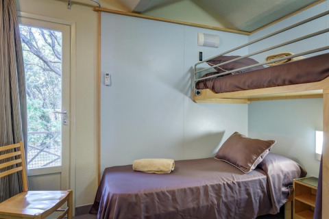 Mobile home bedroom with a single bed, bunk bed, brown bedding, and window overlooking trees.