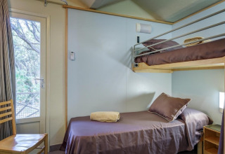 Mobile home bedroom with a single bed, bunk bed, brown bedding, and window overlooking trees.