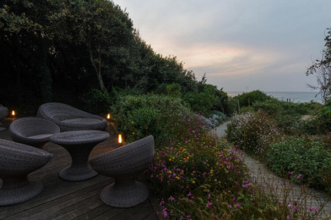 Outdoor patio with wicker furniture, lush garden, and path leading to the sea at sunset in Italy.