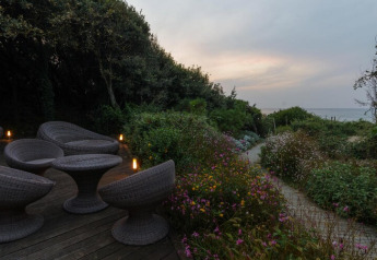 Outdoor patio with wicker furniture, lush garden, and path leading to the sea at sunset in Italy.