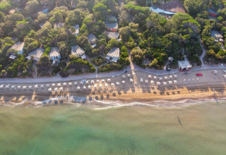 Aerial view of a mobile home site by the beach with umbrellas, cabins, and lush green trees behind.
