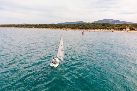 A person is sailing a small boat near the shore with mountains and trees visible behind a mobile home site.