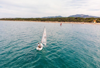 A person is sailing a small boat near the shore with mountains and trees visible behind a mobile home site.