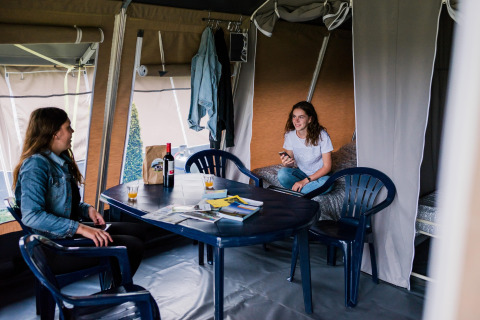 Twee vrouwen zitten te praten aan een tafel in een safaritent met drankjes op tafel.
