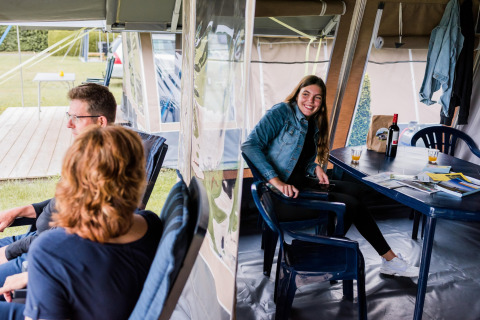 Trois personnes se détendent dans une tente safari, une femme sourit à table entourée d’affaires de camping.