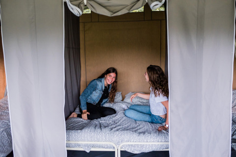 Deux femmes assises sourient sur un lit à l'intérieur d'une tente safari, rideaux tirés sur les côtés.