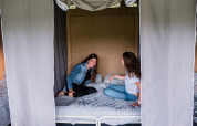 Deux femmes assises sourient sur un lit à l'intérieur d'une tente safari, rideaux tirés sur les côtés.