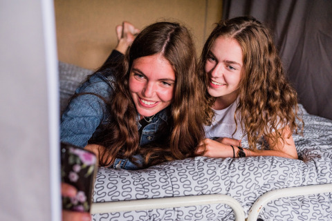 Twee meisjes maken een selfie op een bed in een safaritent, allebei vrolijk lachend naar de camera.