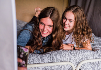 Deux jeunes femmes allongées sur un lit dans une tente safari prennent un selfie en souriant.