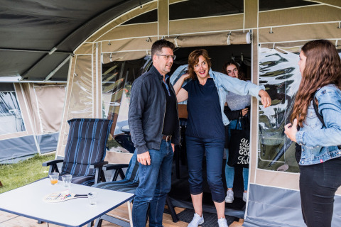 Een familie staat aan de ingang van een safaritent op een camping, lachend en klaar om te ontspannen.