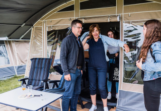 Een familie staat aan de ingang van een safaritent op een camping, lachend en klaar om te ontspannen.