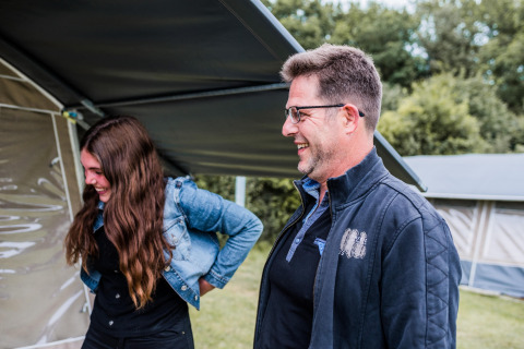 Twee mensen lachen samen onder de luifel van een safaritent op een camping tussen het groen.