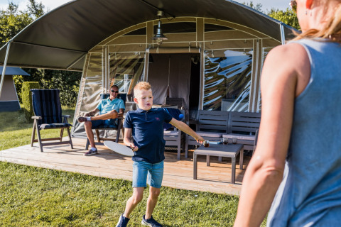 Dreng spiller badminton foran en safaritelt, mens en voksen og endnu en person slapper af på terrassen.